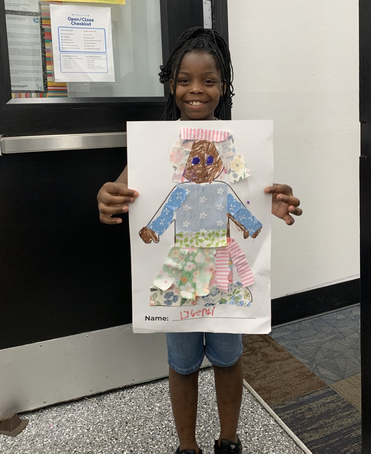 A young girl stands indoors, smiling and holding up a handmade artwork of a person, labeled with the name "Deeni.