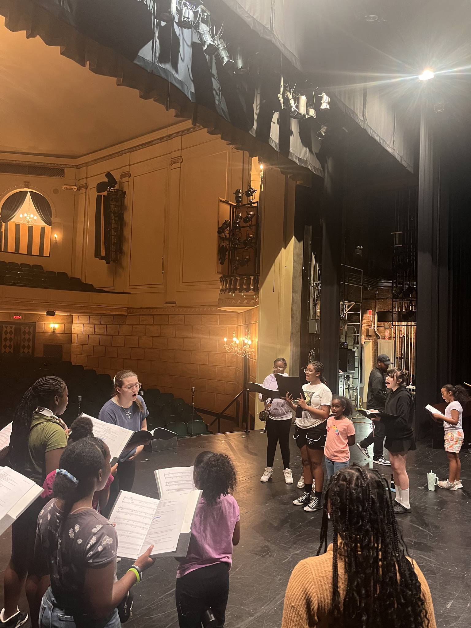 A group of people on a stage rehearsing with music stands and sheet music in a theater, some standing and some seated. The auditorium seats are visible in the background.