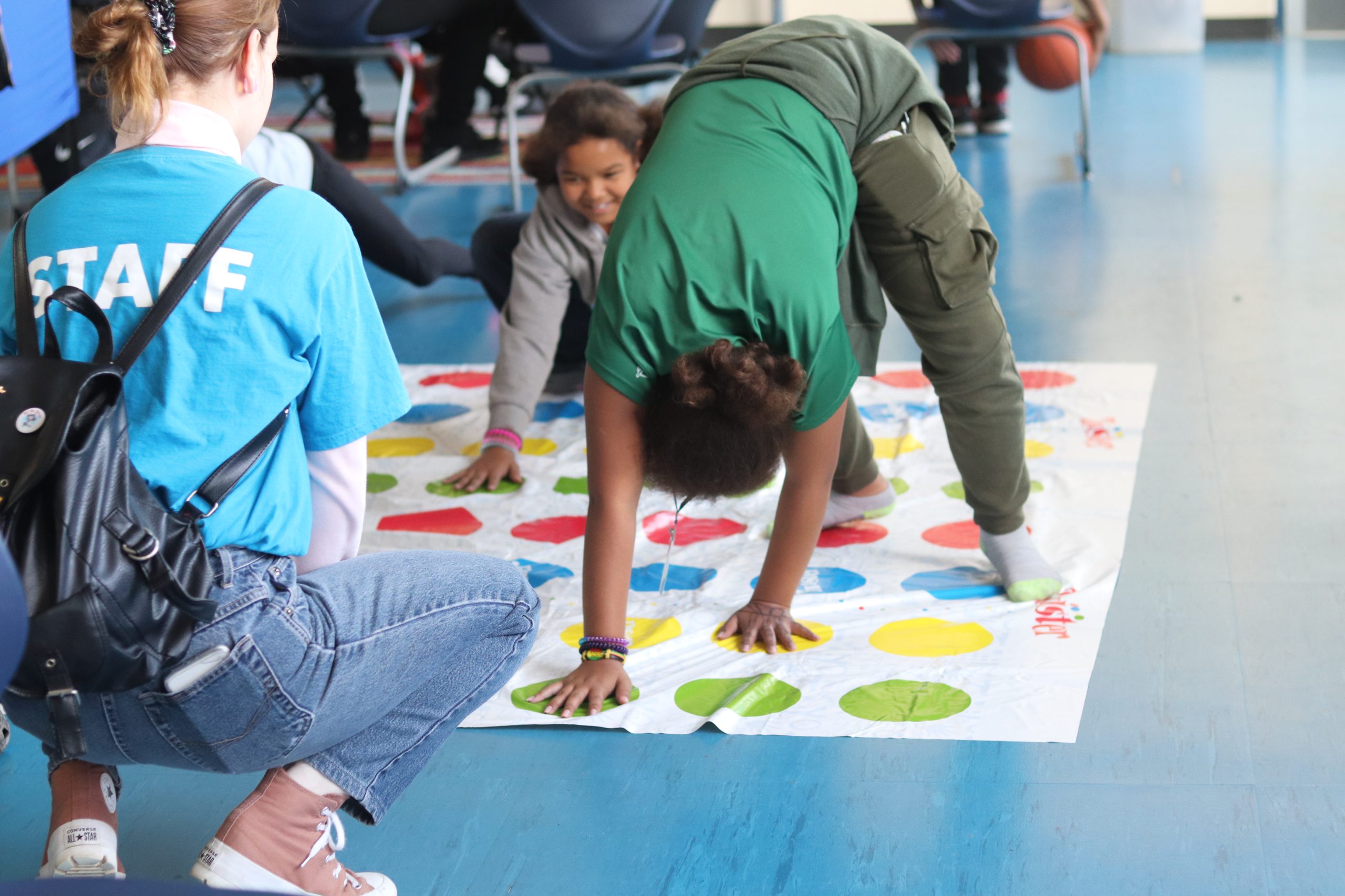 Two children play Twister on a mat with colored circles, supervised by a staff member in a blue T-shirt and jeans.