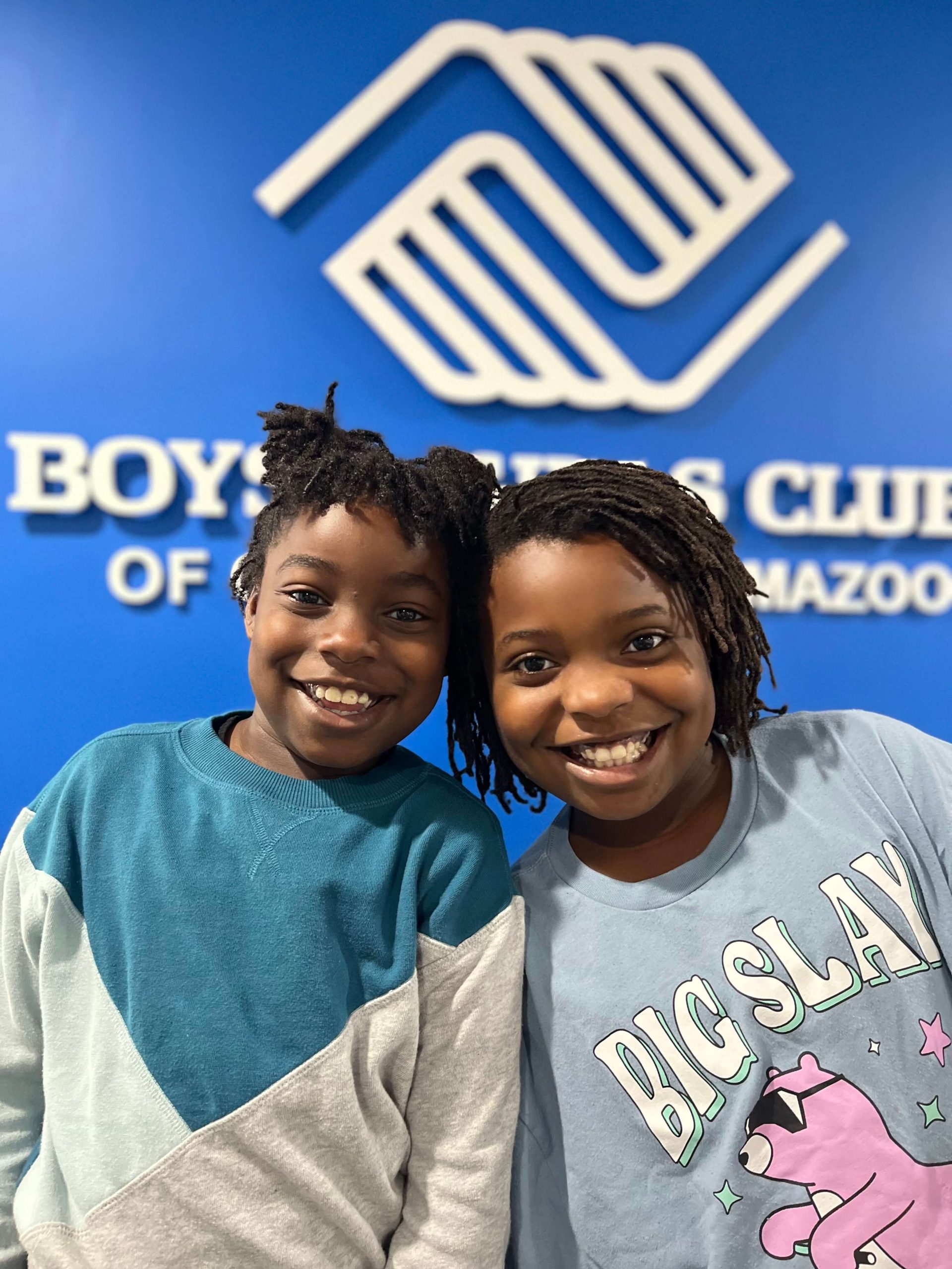 Two smiling children stand in front of a blue wall with the Boys & Girls Club of Kalamazoo logo and text.
