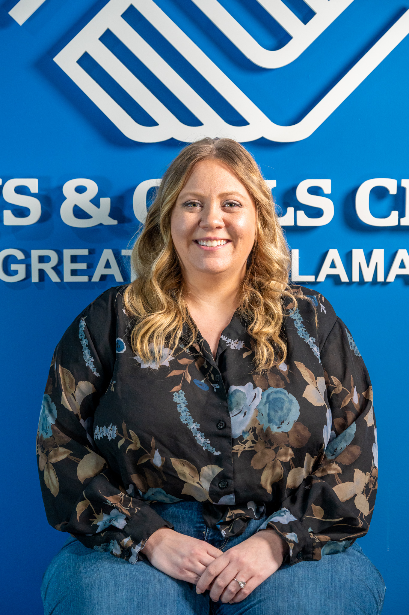 A woman with long blonde hair wearing a floral blouse sits and smiles in front of a blue wall with a Boys & Girls Club logo and text.