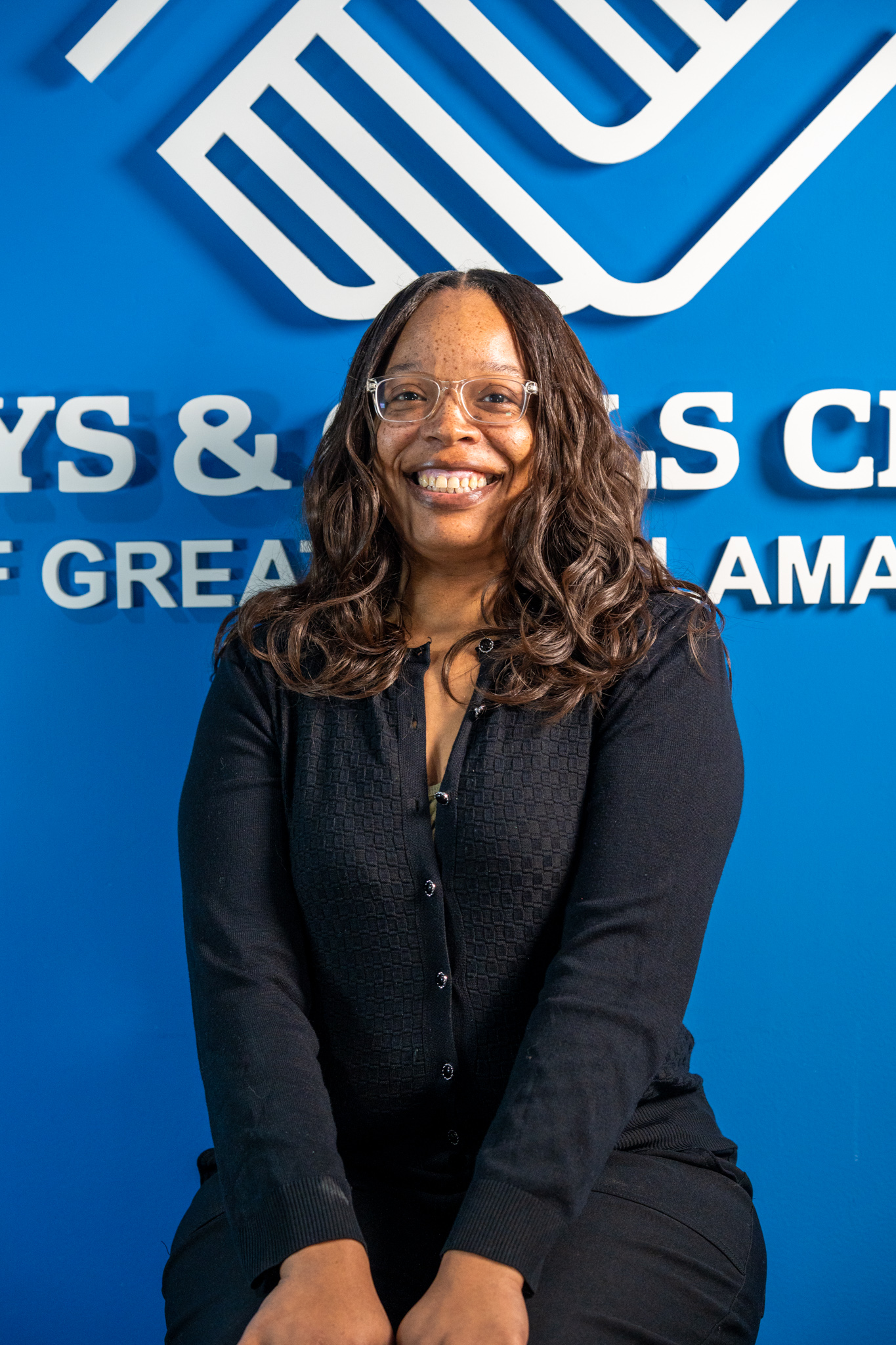 A woman with glasses and long curly hair smiles while seated in front of a blue Boys & Girls Club sign.