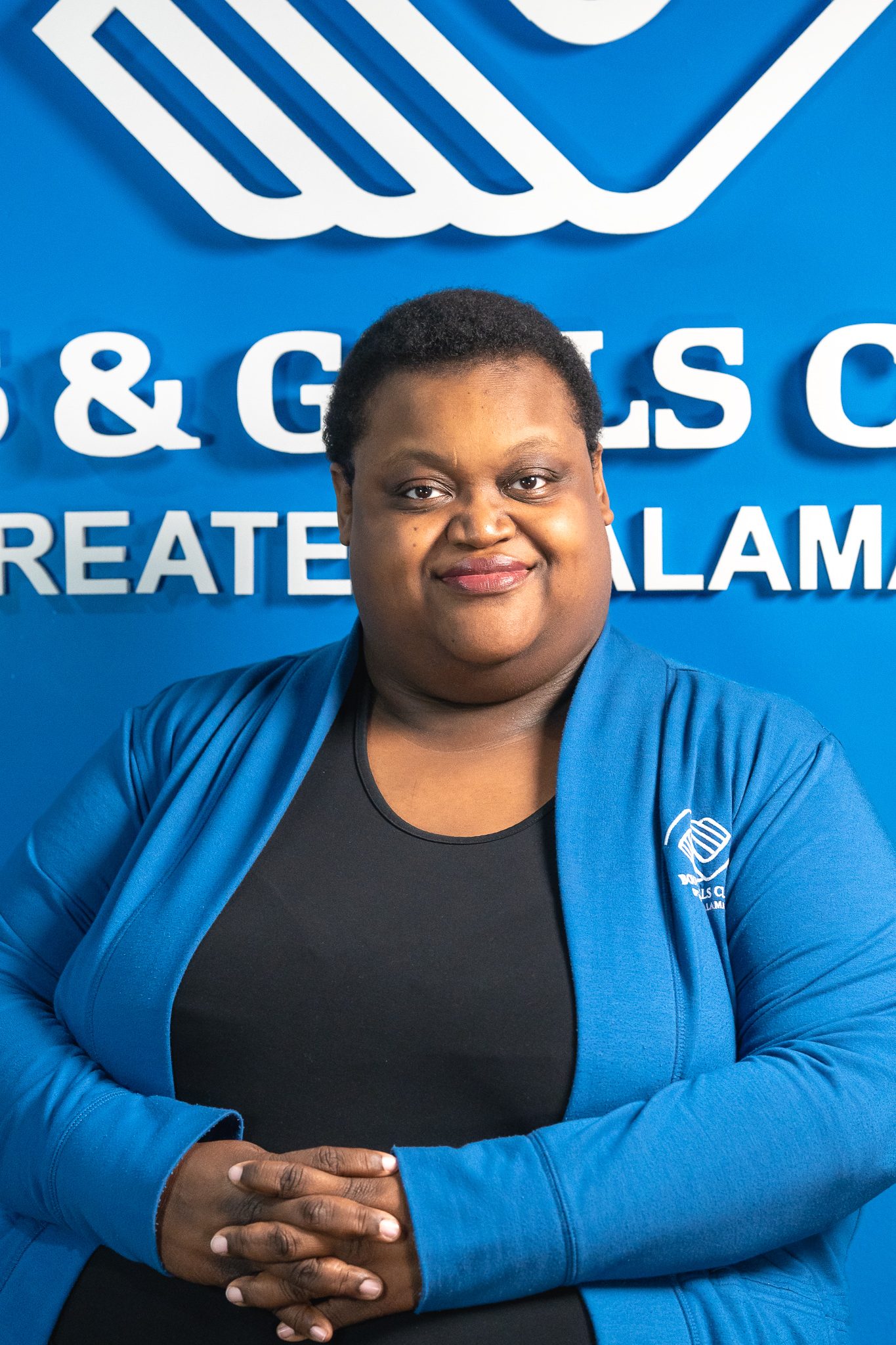 A person wearing a blue jacket sits with hands folded in front of a blue wall displaying the Boys & Girls Club logo and partial text.