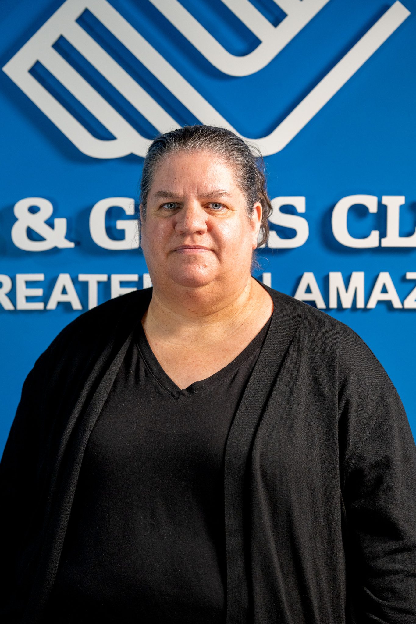 A woman with gray-streaked hair stands in front of a blue wall with partially visible Boys & Girls Club signage. She is wearing a black top and cardigan.