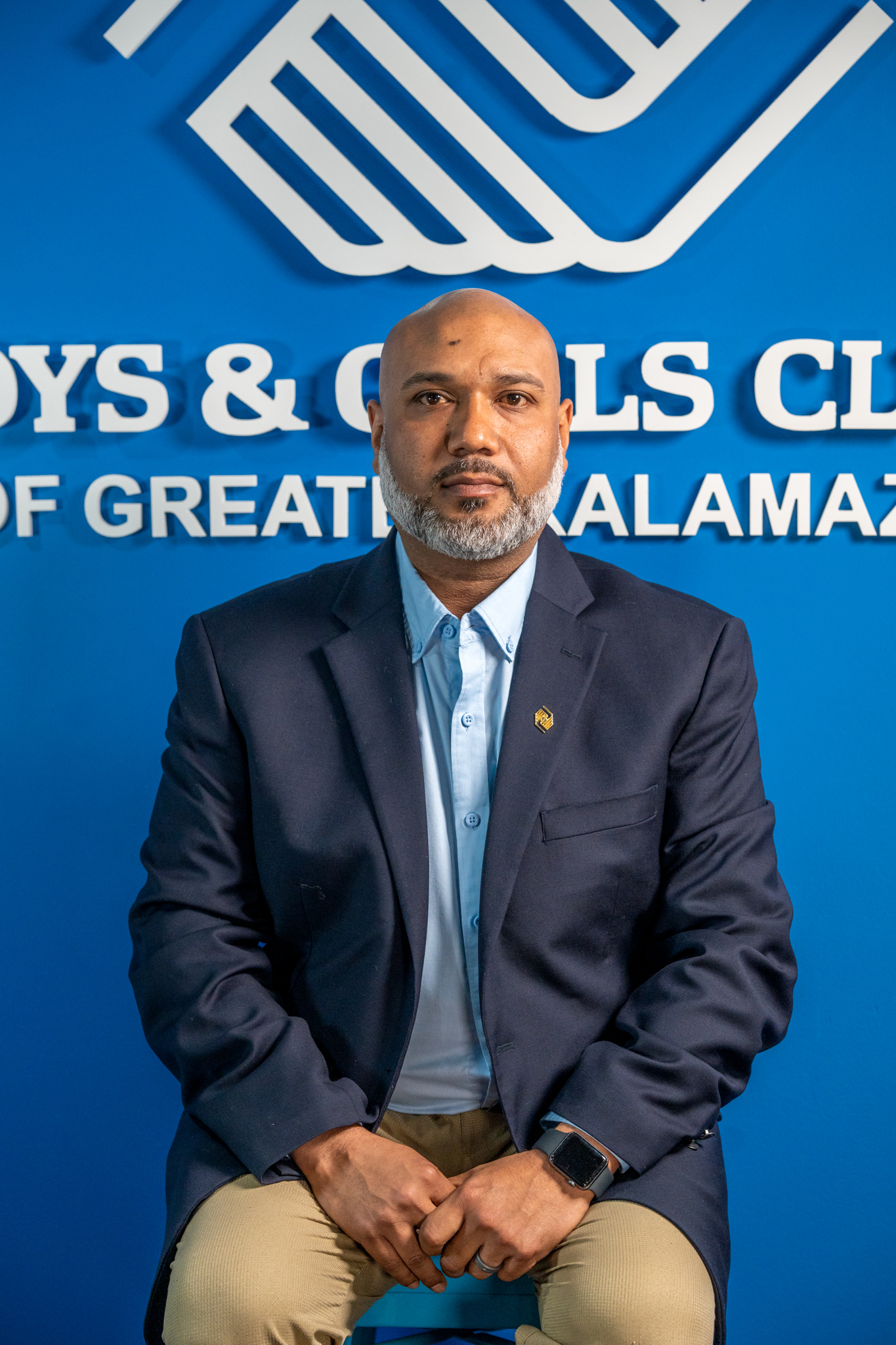 A man in a navy blazer and light blue shirt sits in front of a blue wall with “Boys & Girls Club of Greater Kalamazoo” logo and text.