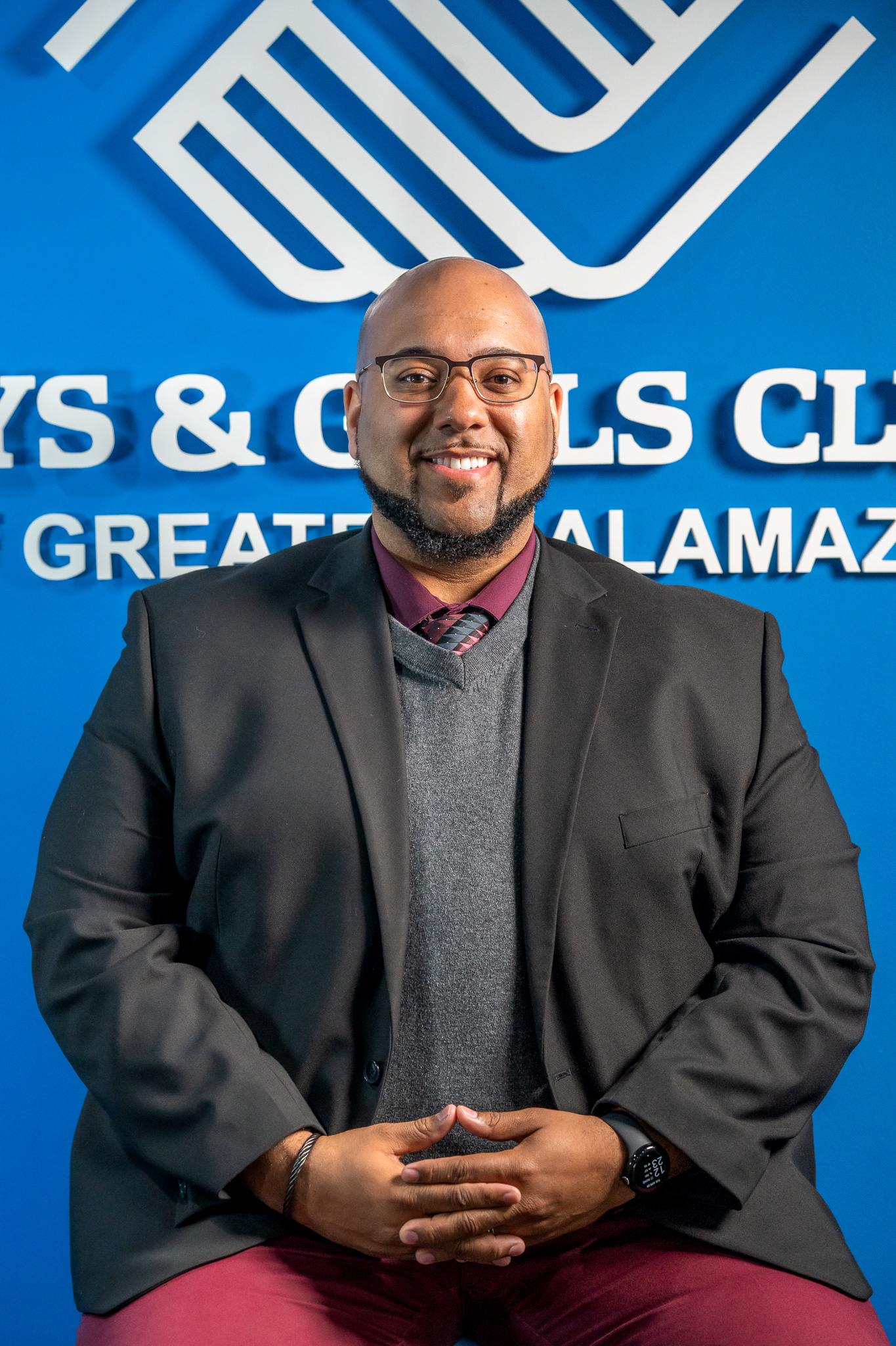 A man in a suit and sweater sits smiling in front of a blue wall with the Boys & Girls Clubs of Greater Kalamazoo logo and text.