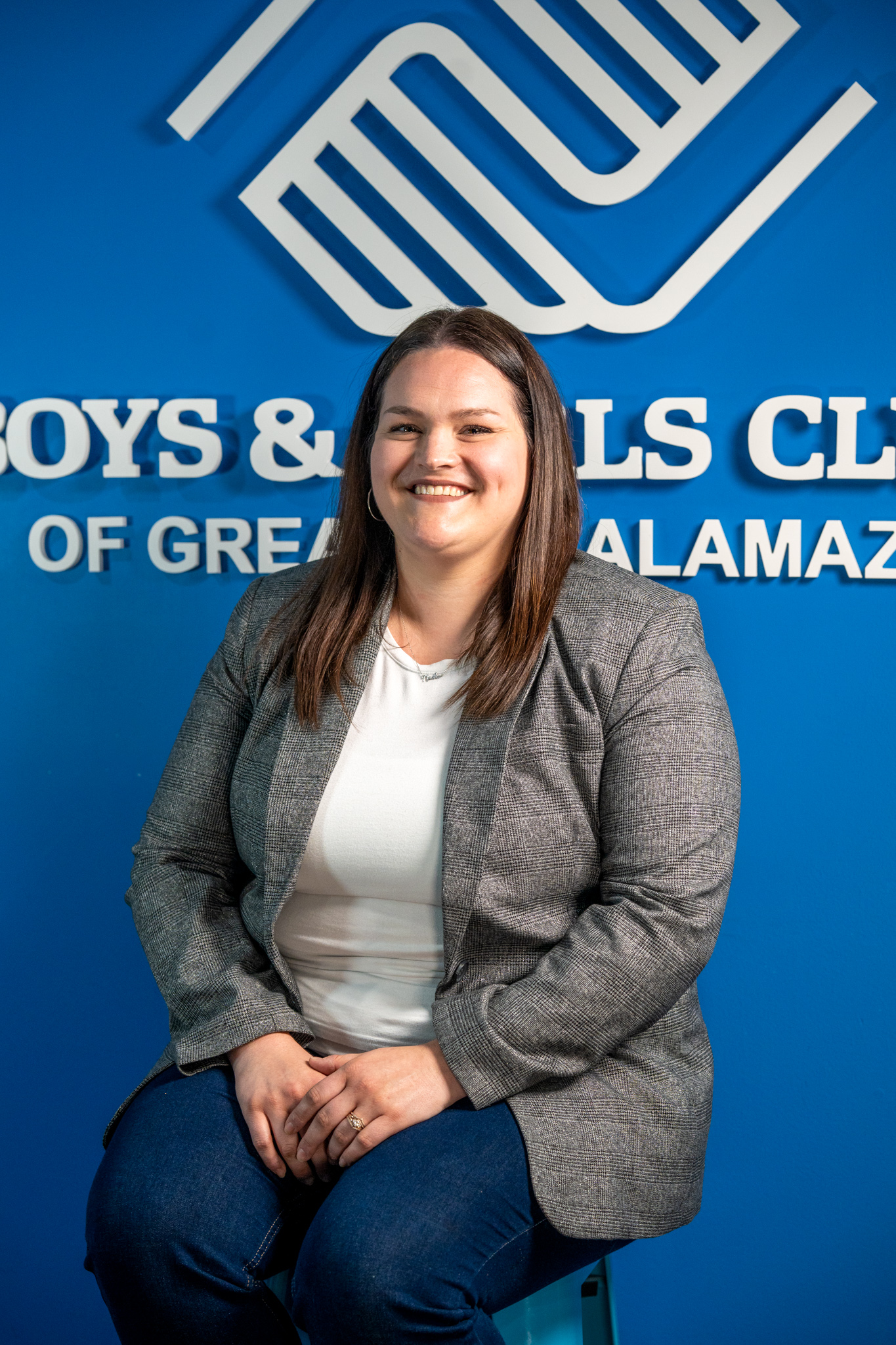 A woman in a gray blazer and white shirt sits and smiles in front of a Boys & Girls Club of Greater Kalamazoo sign on a blue wall.
