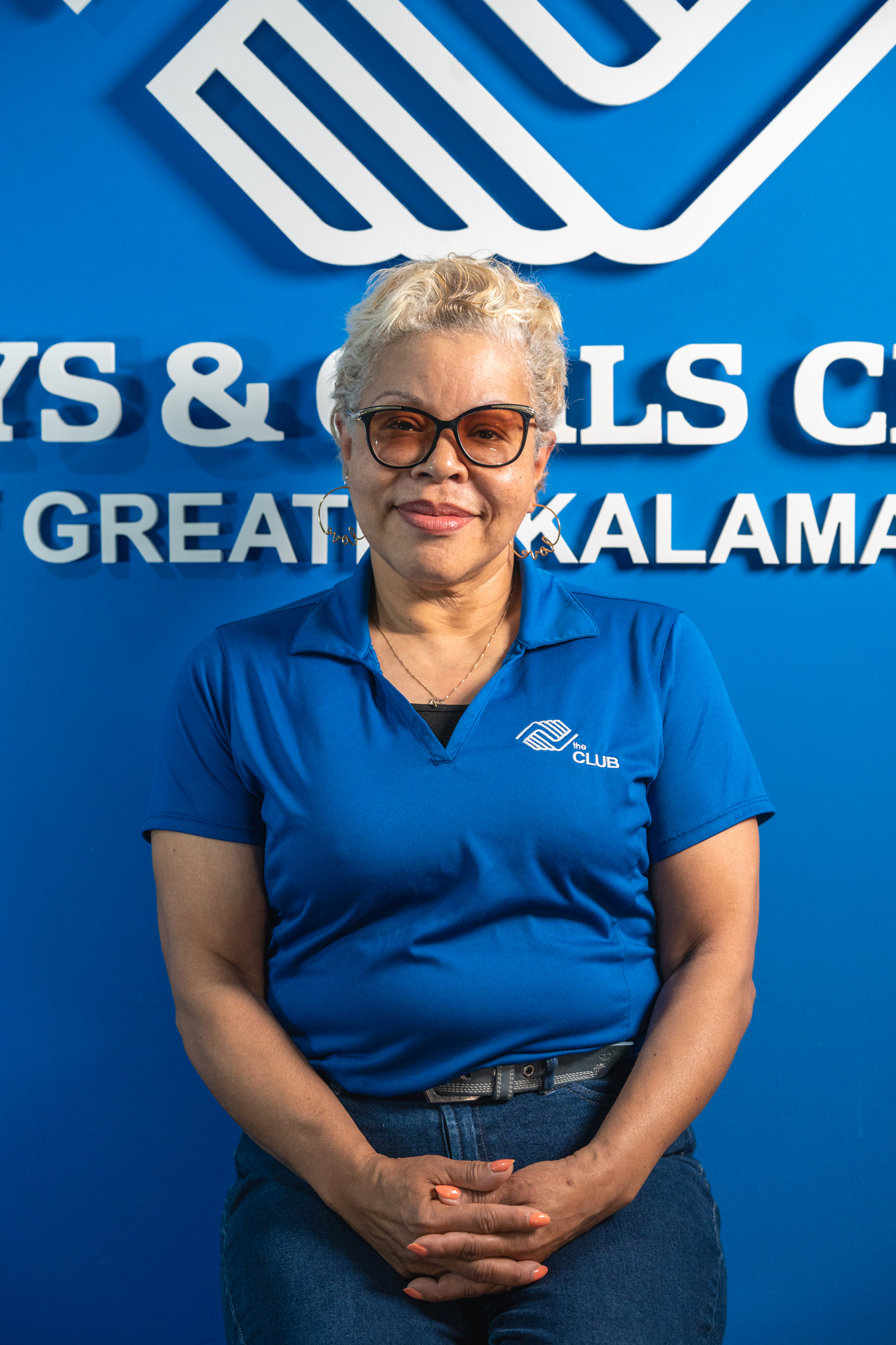 A woman with short gray hair and glasses sits in front of a Boys & Girls Club sign, wearing a blue Club polo shirt and jeans.