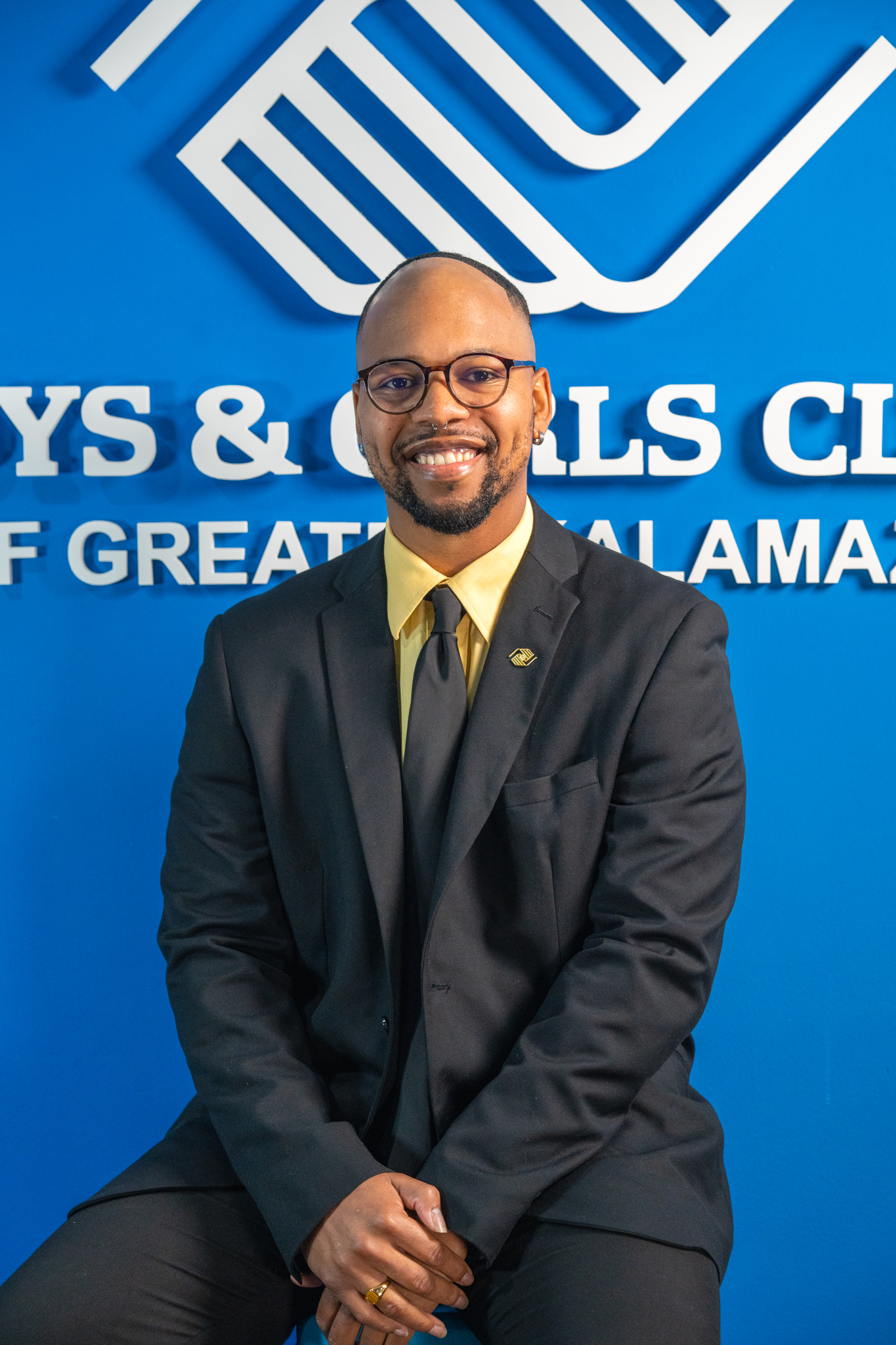 A man in a suit and tie sits and smiles in front of a blue wall with a Boys & Girls Clubs of America logo and partial text in the background.