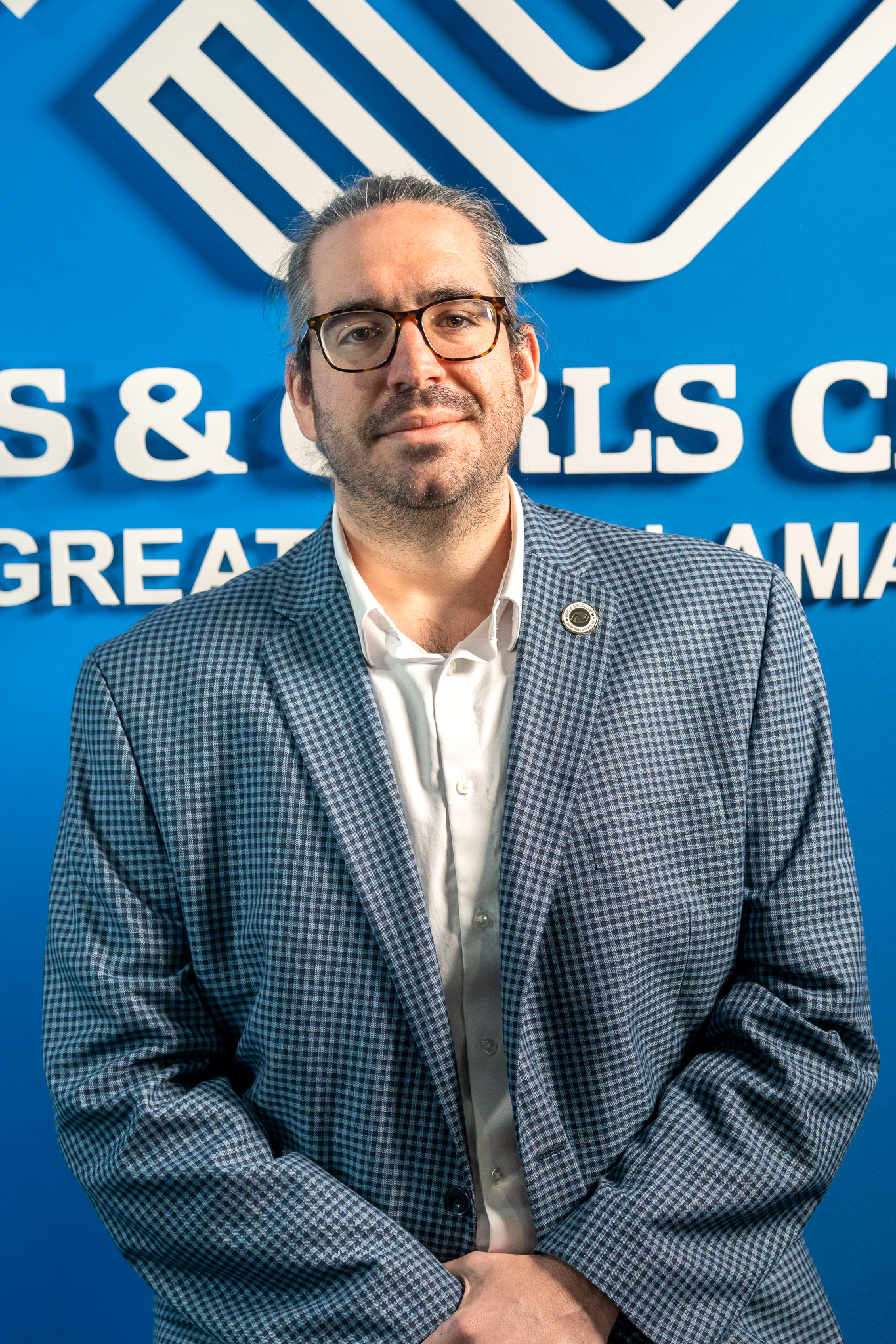 A man wearing glasses and a checked blazer stands in front of a blue wall with partially visible text and a large logo.