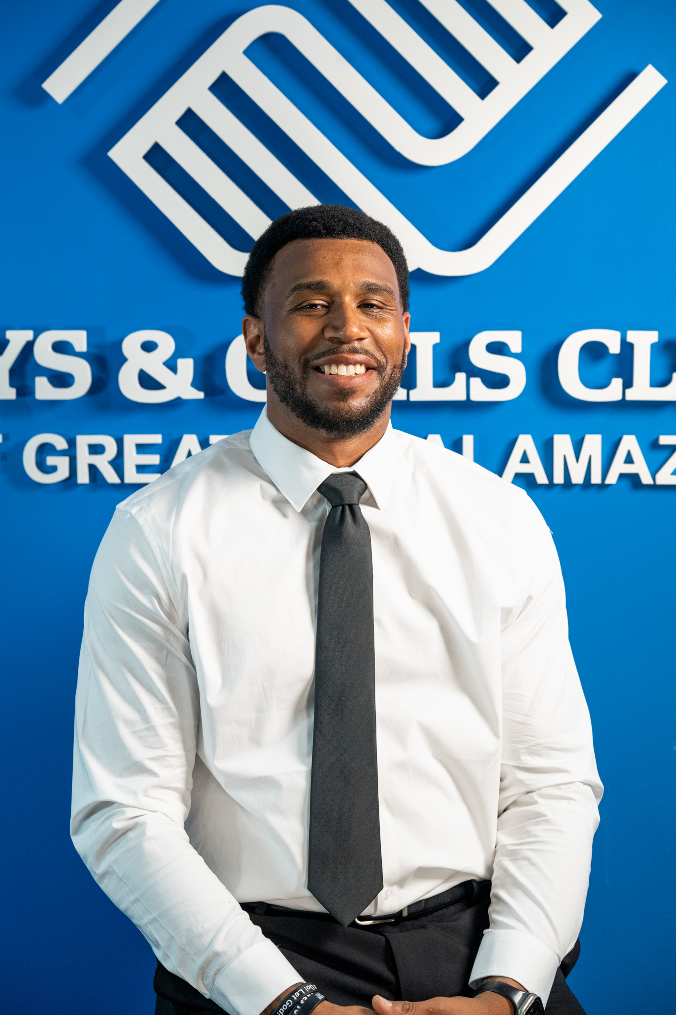 A man in a white shirt and black tie smiles while seated in front of a Boys & Girls Club sign with a blue background.
